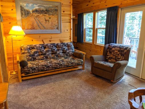 Living room with a futon, chair, and large windows facing the deck and woods.